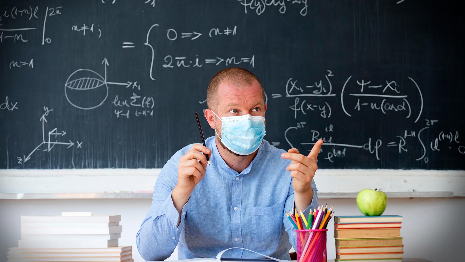 Teacher sitting in front of blackboard with mask on